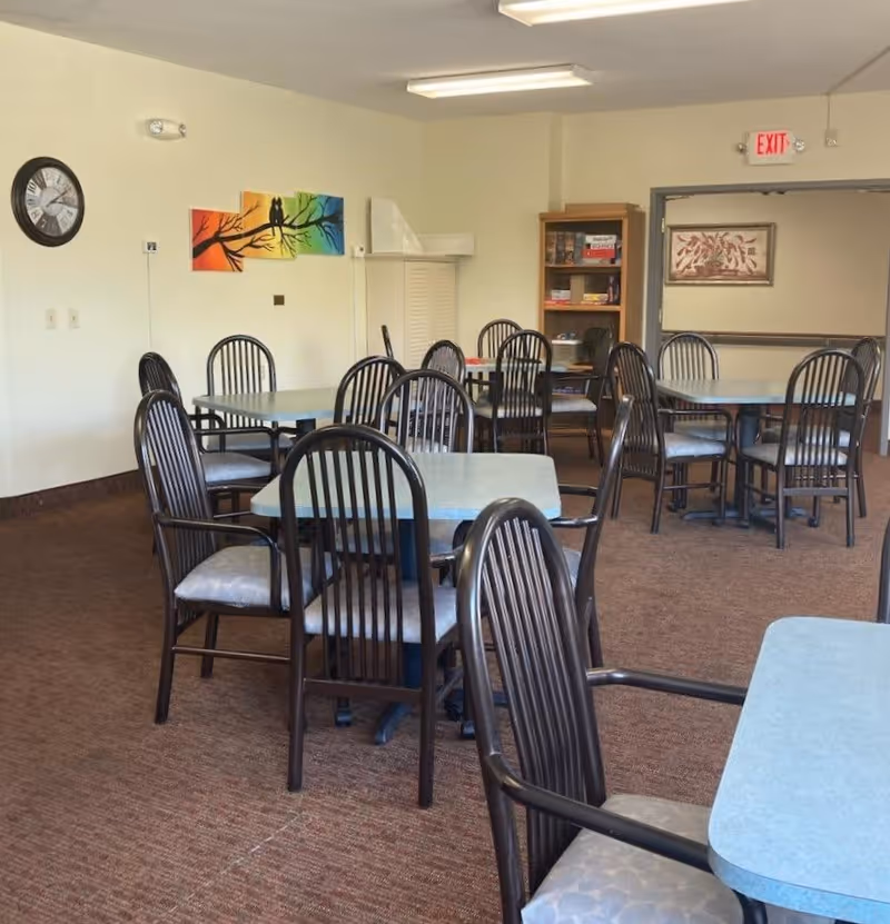 A dining room with multiple square tables and black chairs with cushioned seats. The room has beige walls, a brown carpet, a clock on the wall, colorful artwork featuring a tree branch with two birds, and a bookshelf with games and books. There is an exit sign above a doorway in the background.