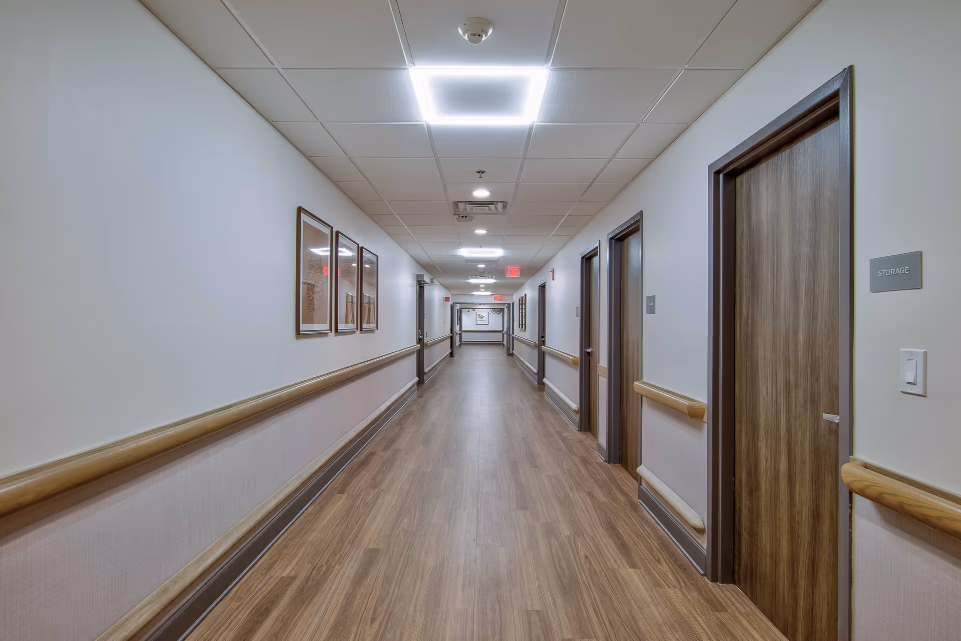 A long, well-lit hallway in a senior living facility with wood flooring, white walls, handrails on both sides, and several closed wooden doors. There are framed pictures on the left wall and a sign labeled 'STORAGE' on the right wall near one of the doors.
