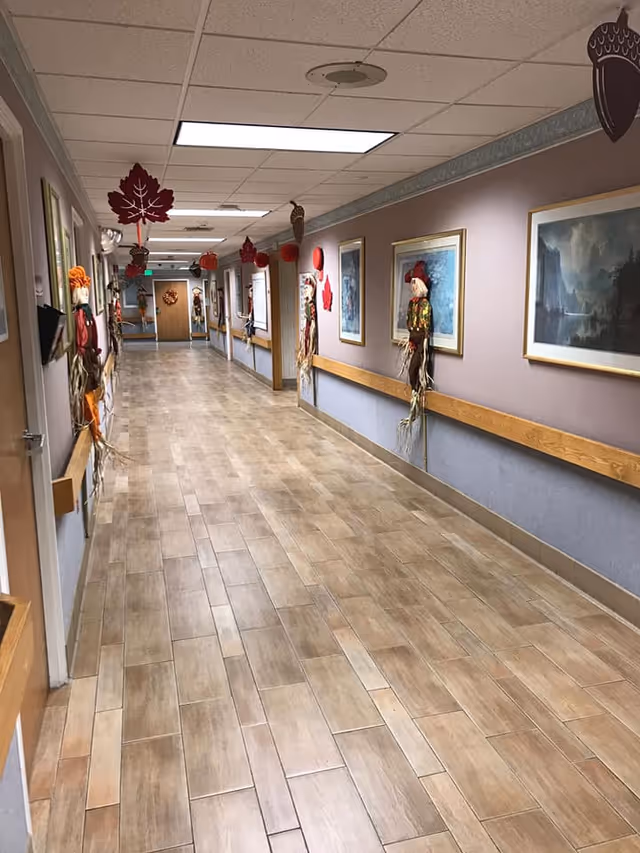 A long, clean hallway in a senior living facility decorated with autumn-themed ornaments including scarecrows, leaves, and acorns hanging from the ceiling and walls. The floor is tiled with wood-like patterns, and framed landscape paintings are hung on the walls. Handrails run along both sides of the hallway.