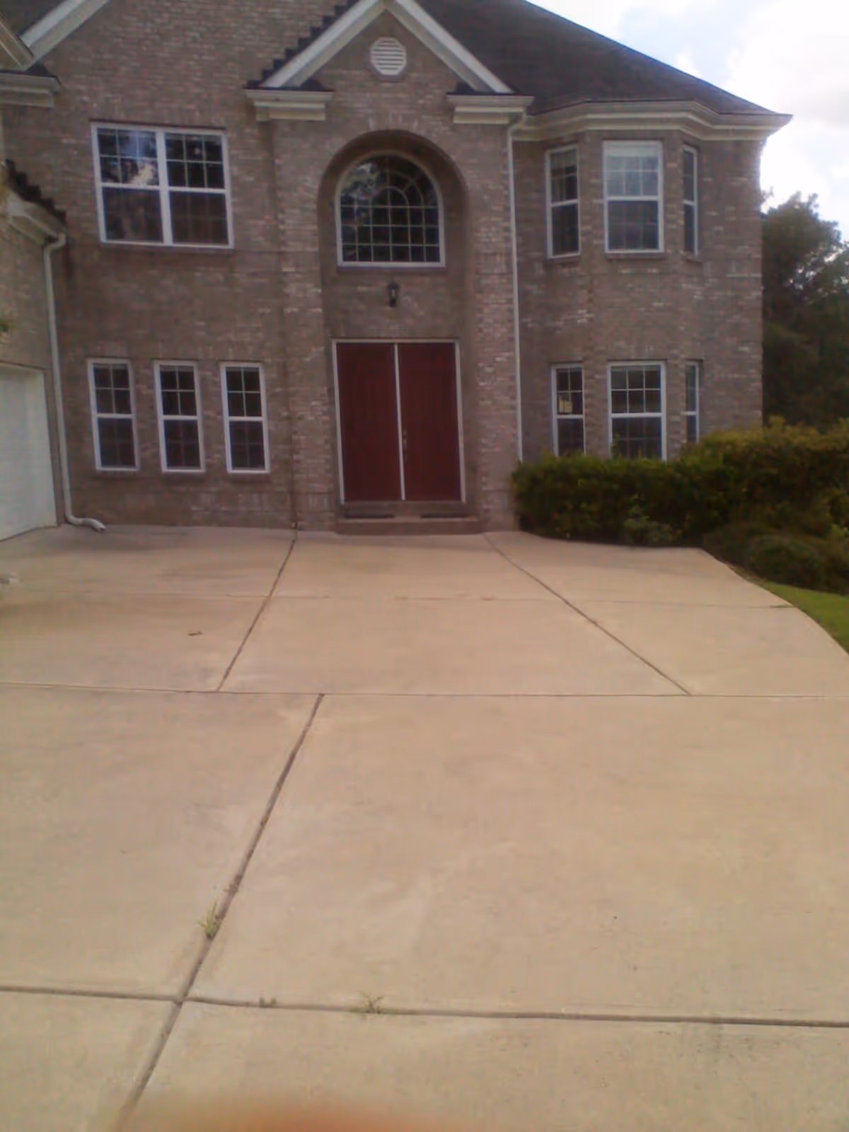 Front exterior view of a two-story brick building with multiple windows, a large arched window above double red doors, and a concrete driveway leading up to the entrance. There are bushes on the right side of the building.