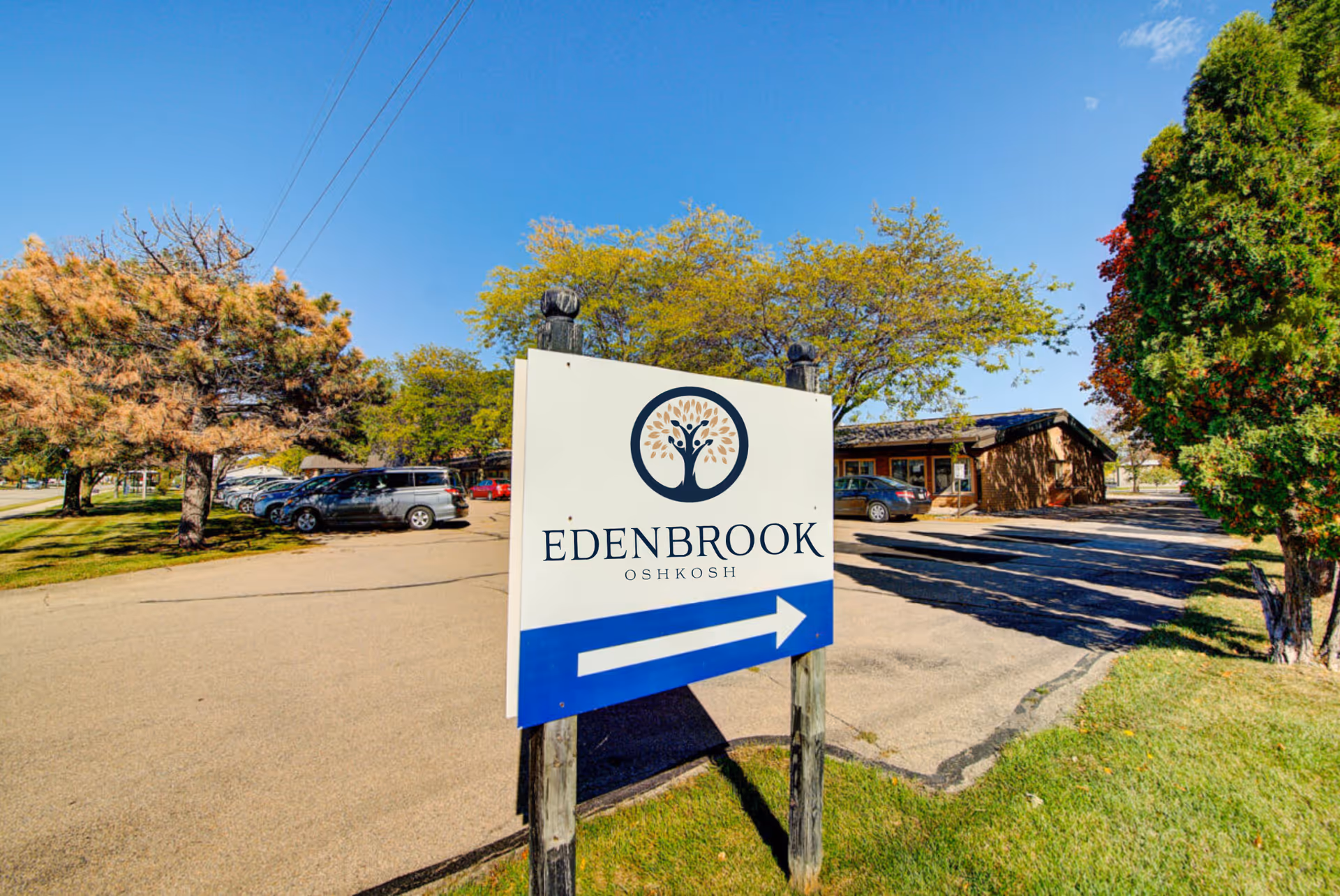 Outdoor view of the Edenbrook Oshkosh facility sign with a blue arrow pointing right, surrounded by trees, parked cars, and a clear blue sky.