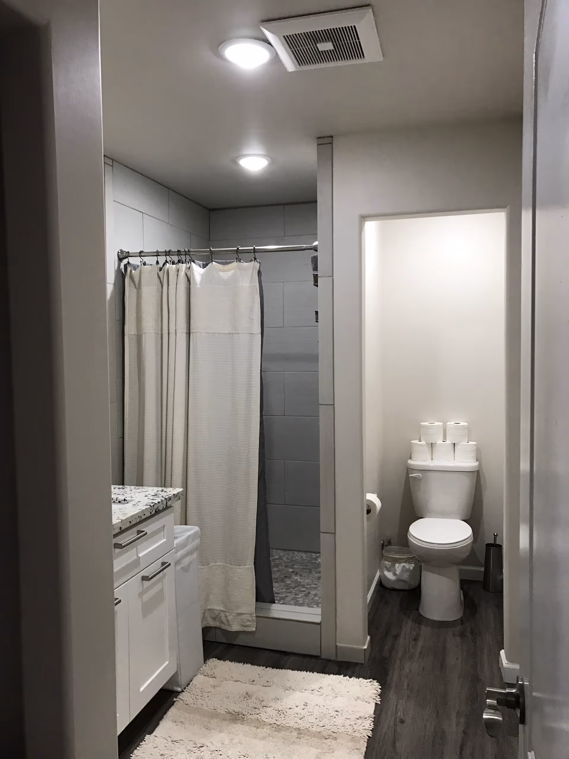 Interior of a bathroom showing a shower with a curtain, a toilet stacked with toilet paper rolls, a white vanity, and a bath mat.