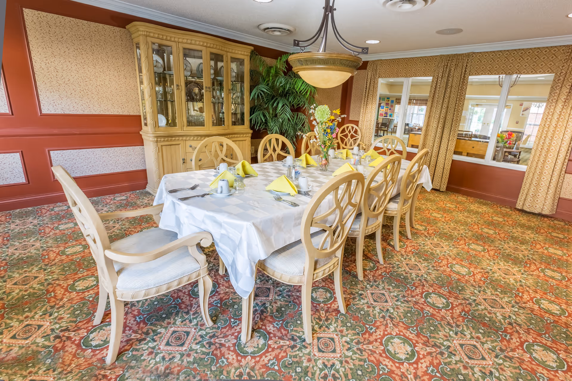 A formal dining room with a long rectangular table covered with a white tablecloth, set with yellow folded napkins, cups, and silverware. The room has patterned carpet, a large wooden china cabinet filled with dishes, a large green plant in the corner, and windows with patterned curtains looking into another room.