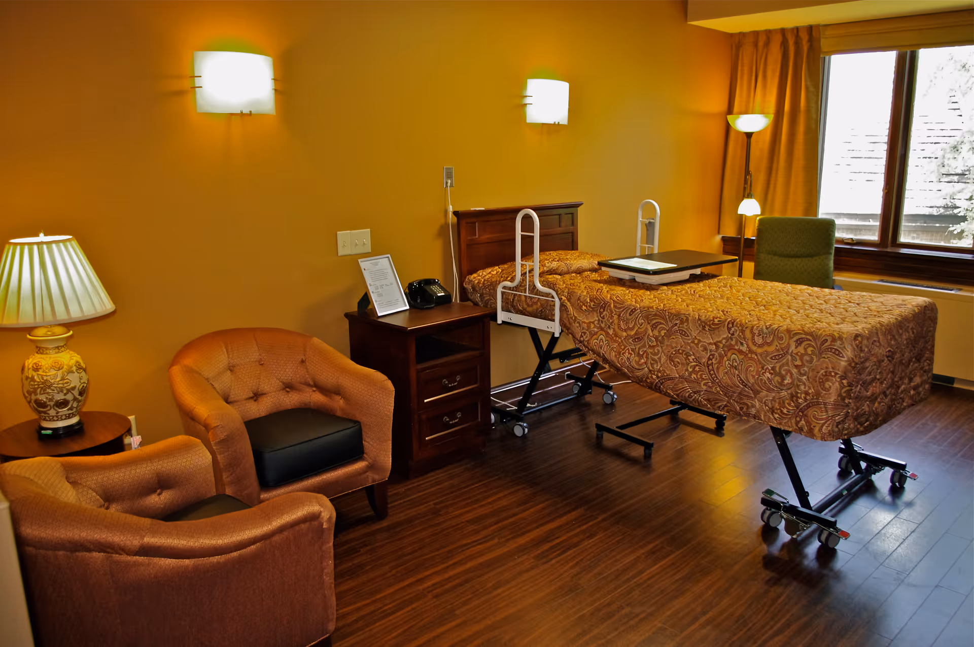 A nursing facility room with a hospital bed covered in a patterned bedspread, two cushioned armchairs, a wooden nightstand with a lamp and telephone, a green armchair near a window with curtains, and warm yellow walls with wall-mounted lights.