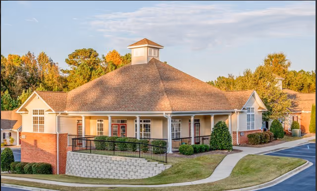 Single-story senior living building with a large hip roof and cupola, a wraparound porch with ramp, red entry doors, and landscaped grounds.