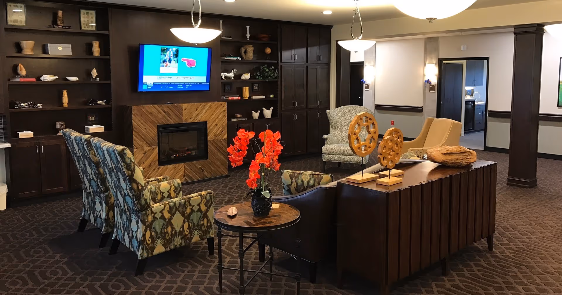 A cozy living room area in a senior living facility featuring patterned armchairs, a small round table with a vase of bright red flowers, a fireplace with a wooden accent, built-in dark wood shelves and cabinets, and a wall-mounted TV. The room has carpeted flooring and soft overhead lighting.