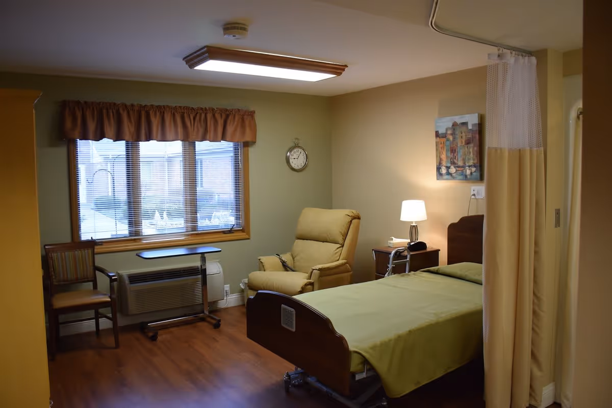 A senior living facility bedroom with a single bed covered in a green blanket, a beige recliner chair, a wooden chair, a small overbed table, a nightstand with a lamp and telephone, a wall clock, and a window with blinds and a brown valance. There is a privacy curtain partially drawn on the right side.