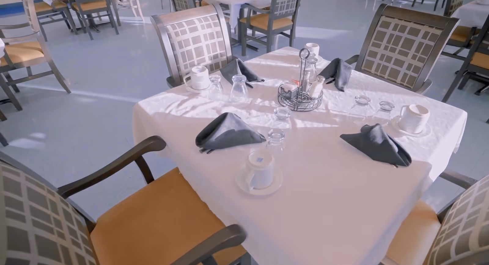 A dining table set for four in a dining room with a white tablecloth, four chairs with patterned backs and tan seats, black folded napkins, cups and saucers, and glassware. Other tables and chairs are visible in the background.