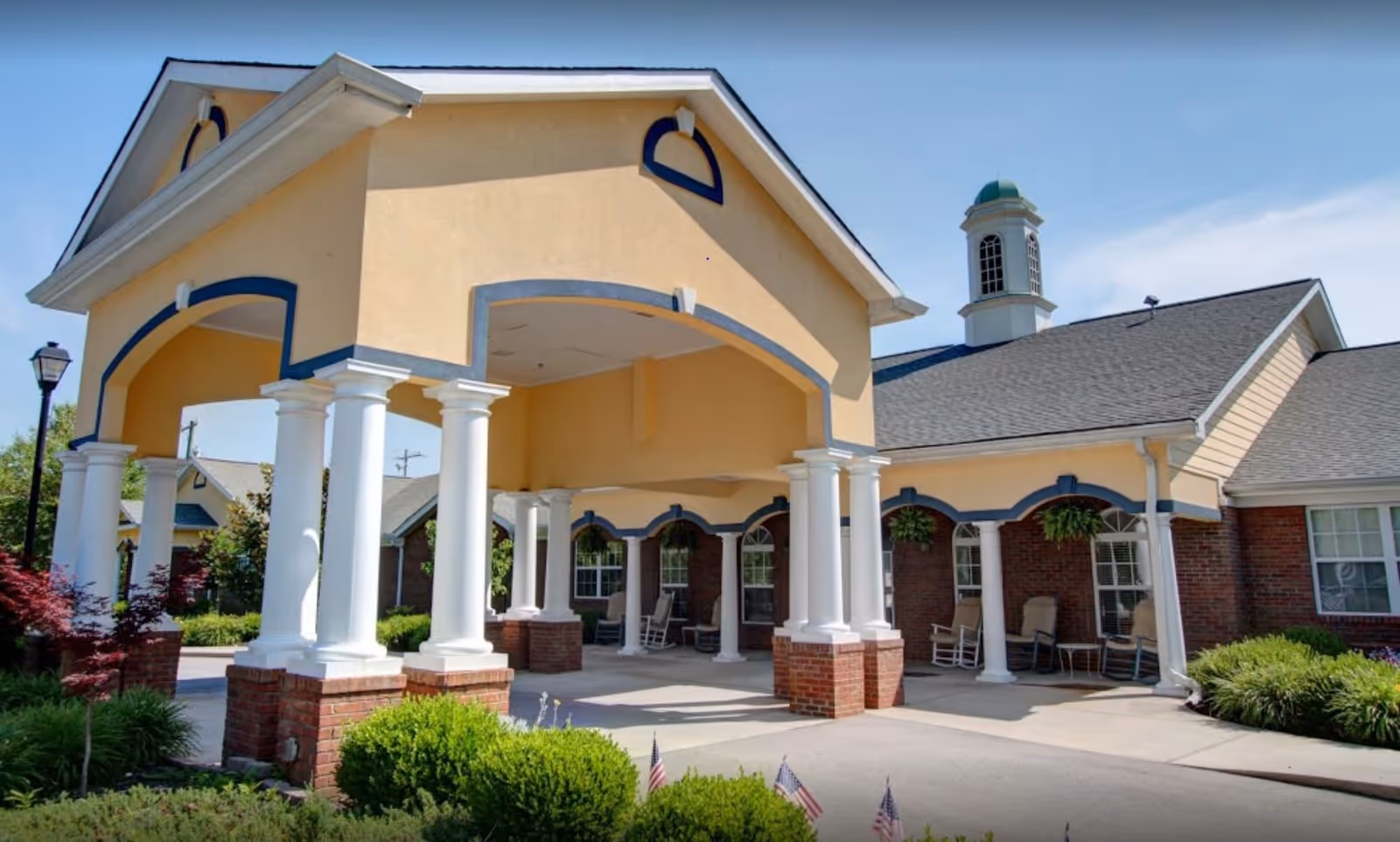 Front exterior view of a senior living facility with a covered entrance supported by white columns on brick bases. The building has a yellow and brick facade with a cupola on the roof and rocking chairs on the porch. There are shrubs and small American flags in the landscaped area near the entrance.
