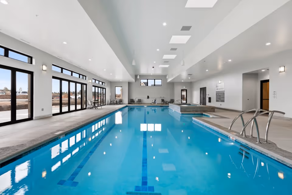 Indoor swimming pool area with clear blue water, surrounded by large windows and glass doors letting in natural light. There are chairs and tables along the sides, and a hot tub adjacent to the pool. The ceiling has recessed lighting and skylights.