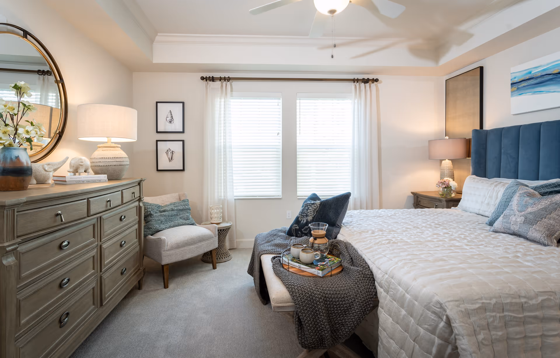 Bright, staged bedroom with a large upholstered bed, bedside tables and lamps, a dresser with mirror, and an armchair by the window.