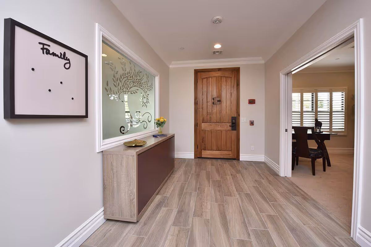 Entryway of a senior living facility with a wooden front door, light wood flooring, a console table with a small vase of yellow flowers, and a decorative frosted glass window with a tree design. To the right, there is an open doorway leading to a room with a dining table and chairs, and windows with white shutters. A wall decoration with the word 'Family' is mounted on the left wall.