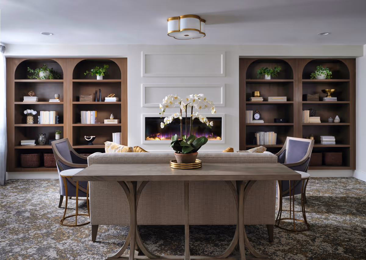 A cozy living room with a beige sofa facing a modern electric fireplace set in a white paneled wall. On either side of the fireplace are built-in dark wood shelves filled with books, plants, and decorative items. Two blue armchairs with gold legs flank the sofa, and a wooden console table behind the sofa holds a potted white orchid. The room has a patterned carpet and a ceiling light fixture with a gold trim.