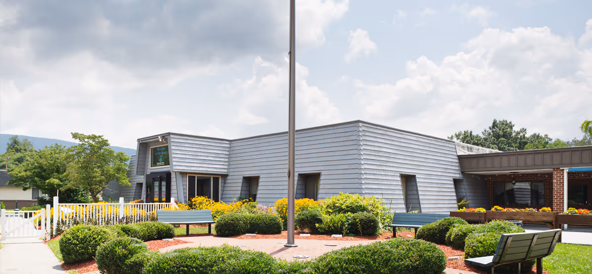 Exterior view of Heritage Hall Rich Creek facility with a modern gray building surrounded by landscaped bushes, yellow flowers, and benches under a partly cloudy sky.