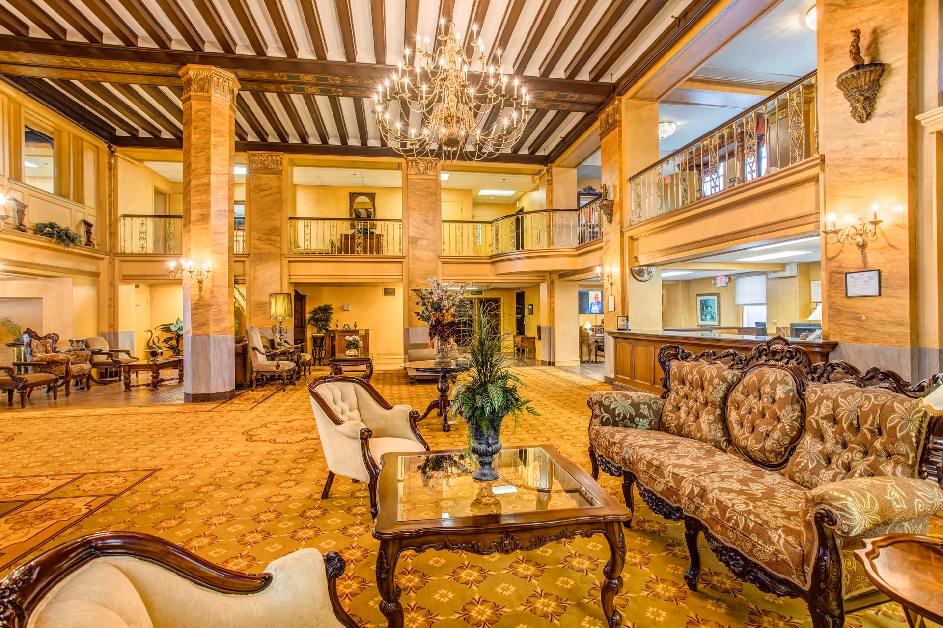A spacious and elegant lobby area with vintage-style furniture including upholstered chairs and a sofa, a glass coffee table with a plant centerpiece, ornate columns, a large chandelier hanging from a beamed ceiling, and a balcony overlooking the lobby. The carpet is patterned and the walls have a warm beige tone with sconces providing soft lighting.