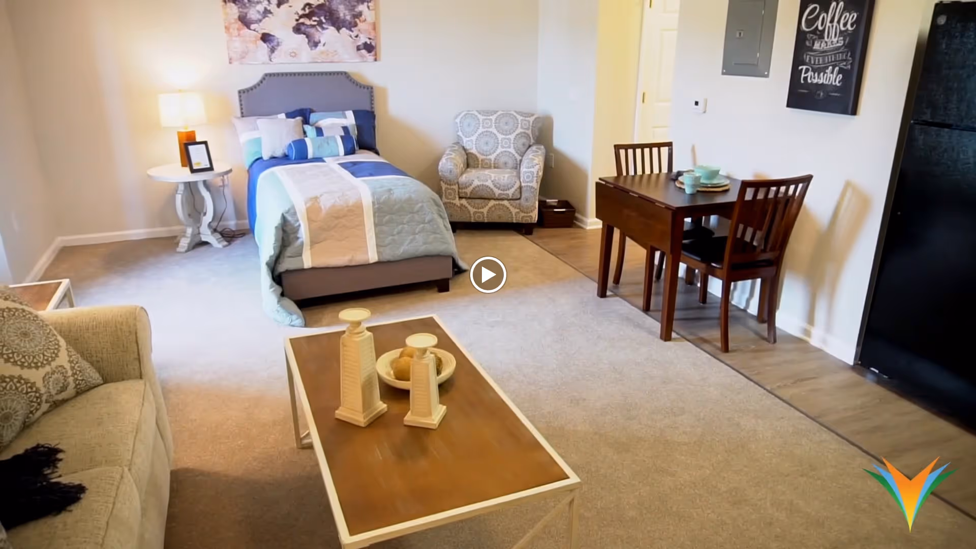 A cozy studio apartment featuring a bed with blue and beige bedding, a patterned armchair, a small wooden dining table with two chairs, a beige sofa, and a wooden coffee table with decorative items. The room has a light carpet and a small kitchen area with a black refrigerator. Wall art and a lamp on a side table add to the homey atmosphere.