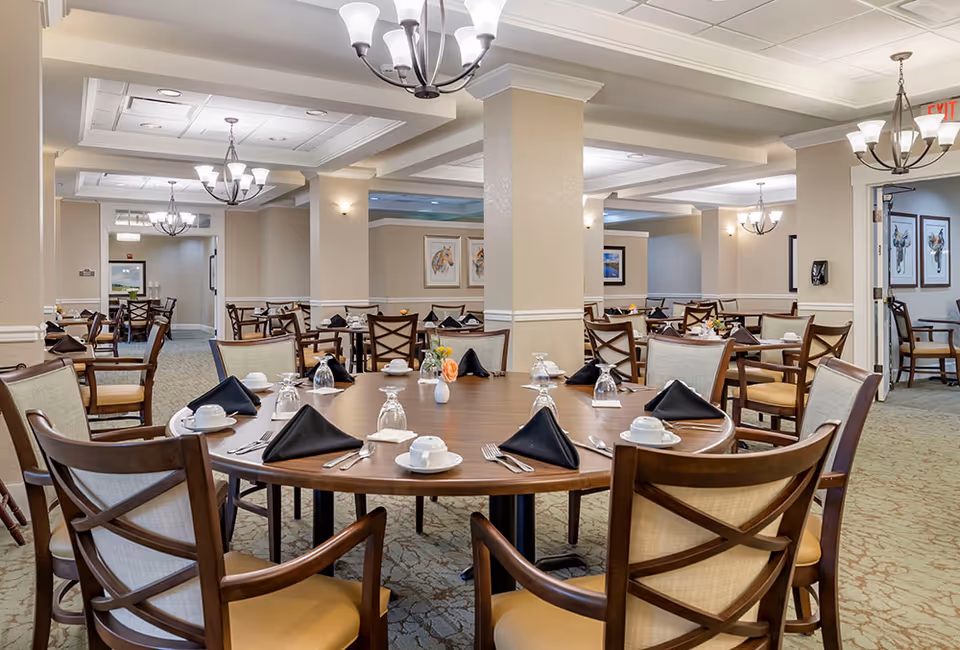 A spacious dining room in a senior living facility with multiple round and rectangular wooden tables set with white cups, saucers, silverware, and black folded napkins. The room features beige walls, carpeted floors, and several chandeliers hanging from the ceiling. Artwork is displayed on the walls, and there are multiple chairs arranged around each table.