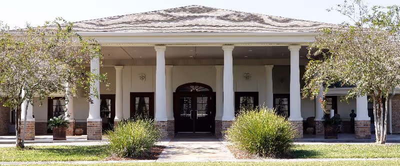 Front exterior view of a building with a covered entrance supported by white columns, surrounded by greenery and small trees.