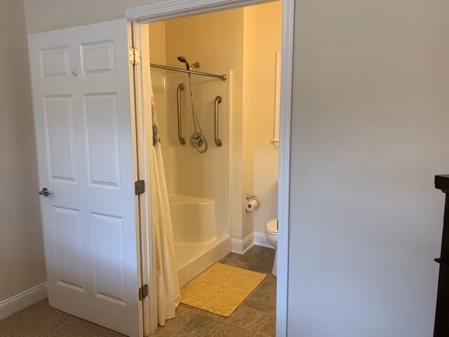 View of a bathroom from an adjacent room, showing a white door partially open. Inside the bathroom, there is a shower with grab bars and a shower curtain, a yellow bath mat on the tiled floor, and a toilet with a toilet paper holder on the wall.