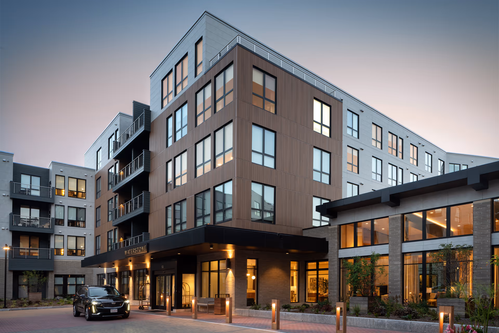 Modern multi-story senior living building entrance at dusk with a covered drop-off canopy reading "WATERSTONE", lit windows, and a car parked in front.