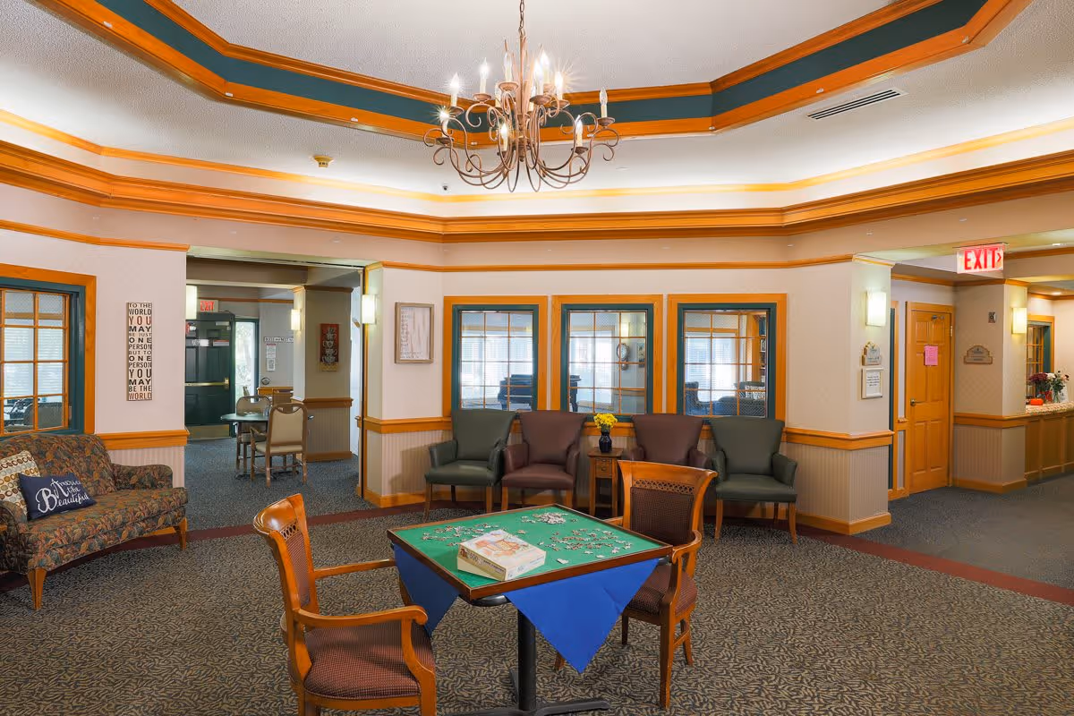 A cozy common area in an assisted living facility featuring a table with a puzzle in progress, surrounded by wooden chairs. The room has a patterned carpet, a chandelier hanging from a tray ceiling with orange trim, and several armchairs against the walls. There are windows with wooden frames and a doorway leading to another room with more seating and tables. The walls are decorated with framed art and a motivational sign.