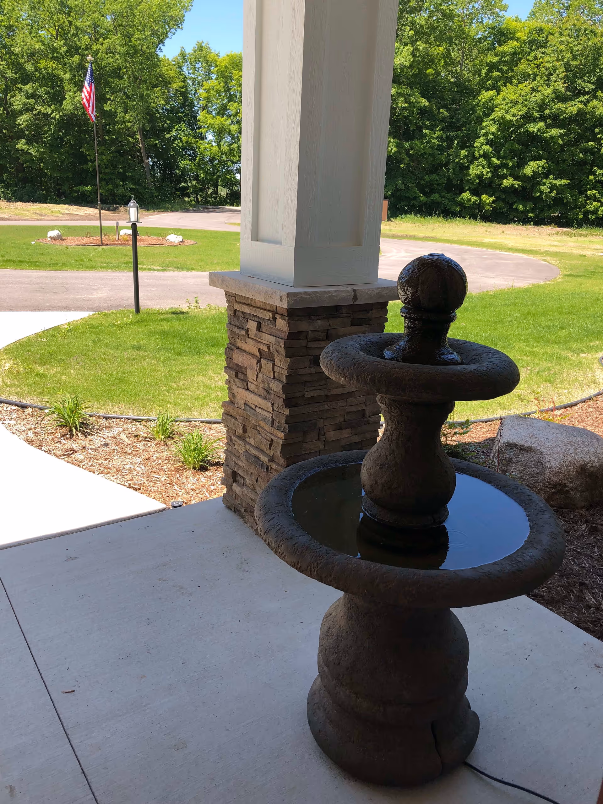 A stone water fountain on a concrete porch with a stone pillar and green lawn in the background. An American flag is visible on a flagpole near a circular driveway, surrounded by trees and landscaping.