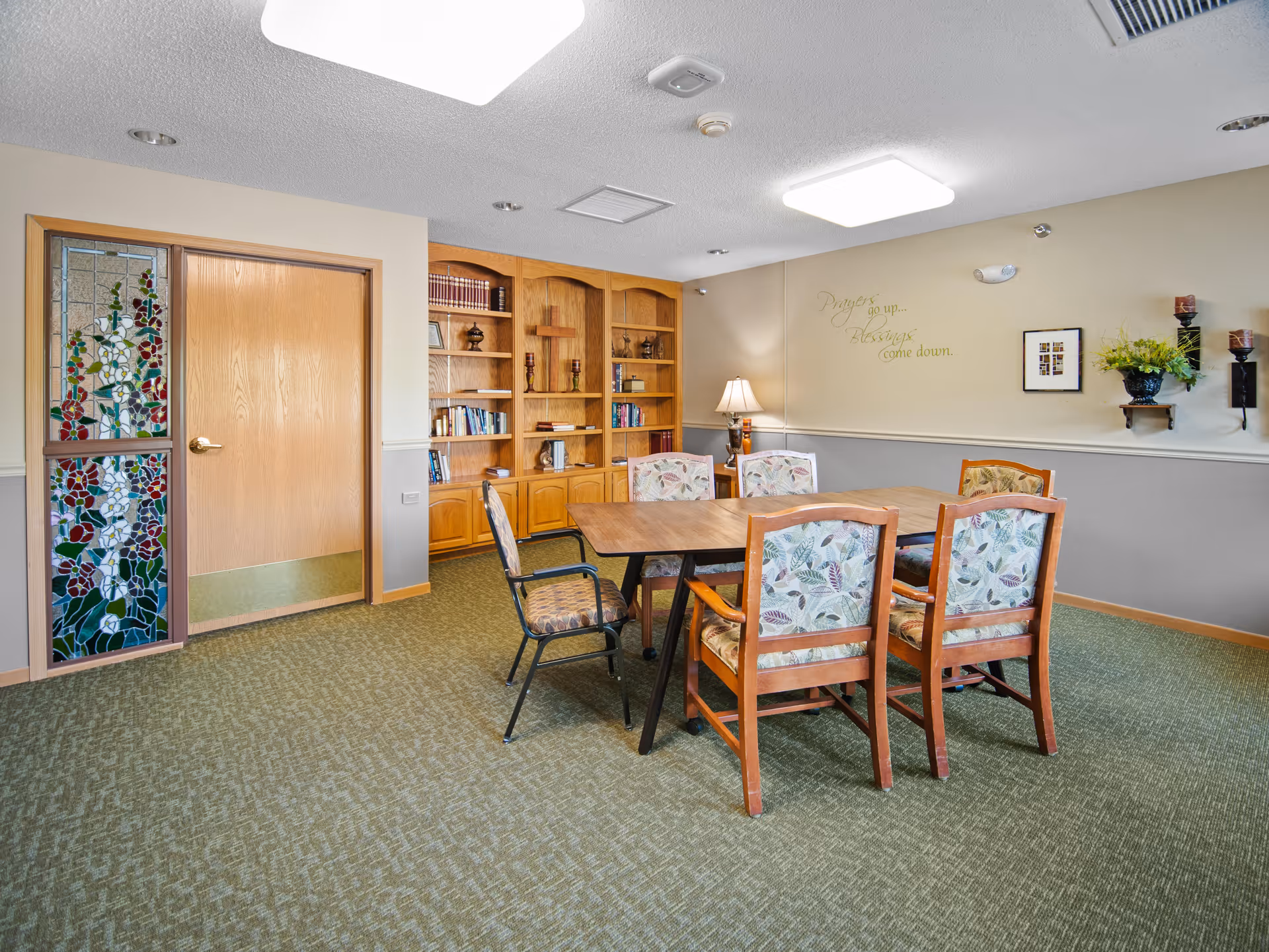 Communal dining/activity room with a wooden table surrounded by floral-upholstered chairs, built-in bookshelves, a stained-glass panel by the door, and wall decor.