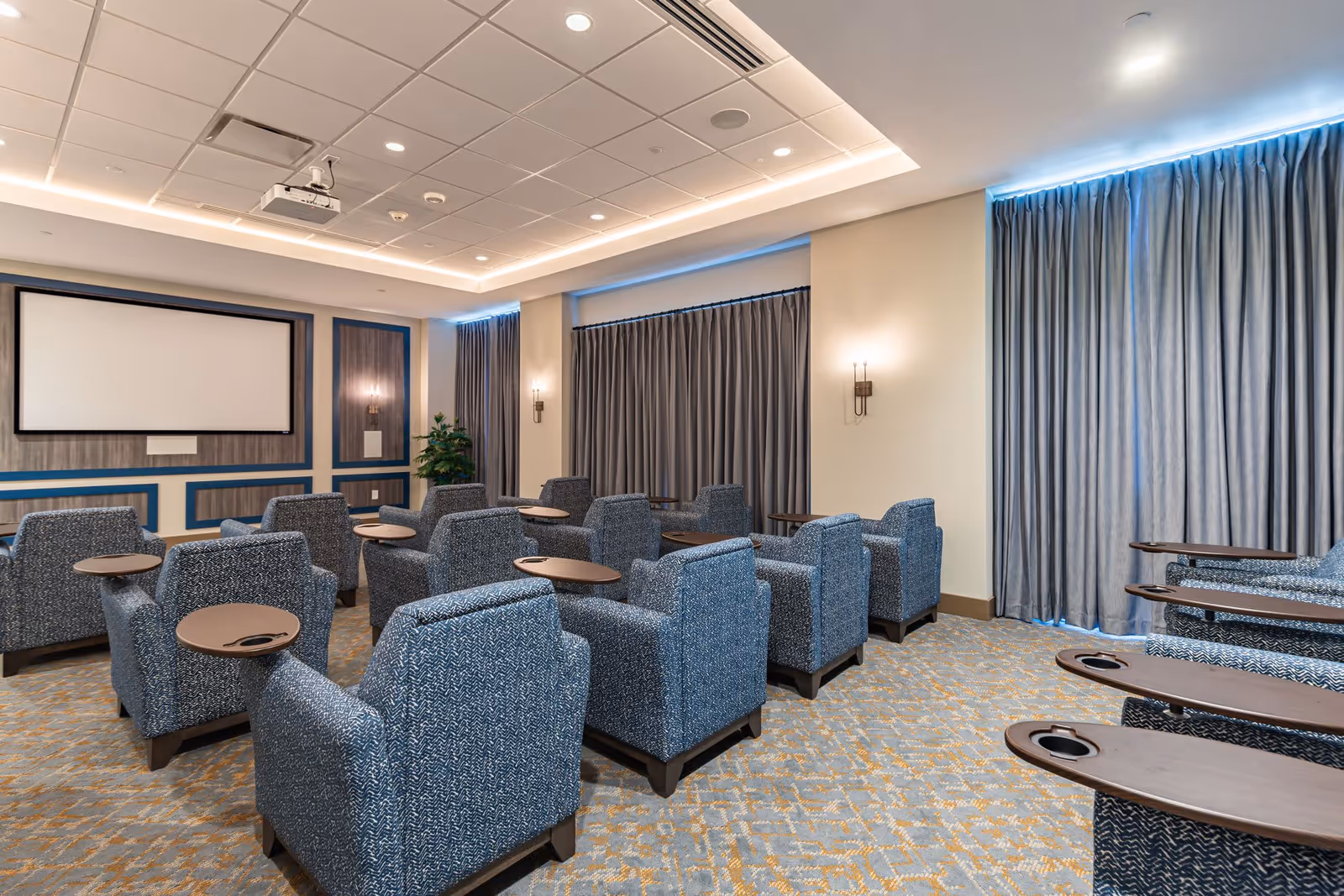 A modern theater room with multiple rows of blue upholstered armchairs, each equipped with a small wooden table with a cup holder. The room features a large projector screen at the front, recessed ceiling lights, wall sconces, and large windows covered with gray curtains.