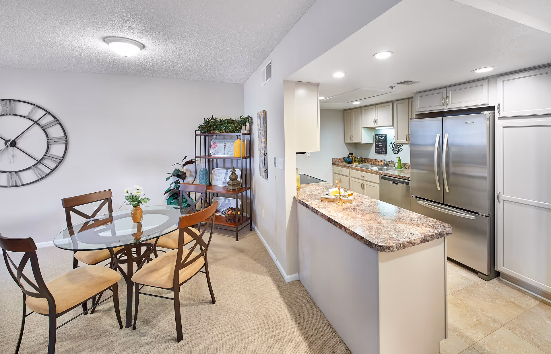 Interior view of a senior living facility dining area and kitchen. The dining area features a round glass table with four wooden chairs with beige cushions. A large decorative wall clock is mounted on the white wall. Next to the dining area is a metal shelving unit with decorative items and plants. The kitchen has white cabinets, granite countertops, a stainless steel refrigerator, dishwasher, and recessed lighting.