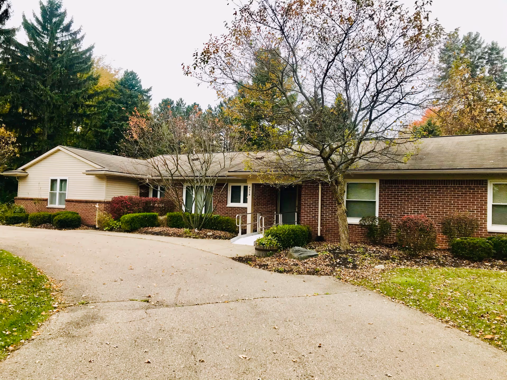 Single-story brick and siding building with a sloped roof, surrounded by trees and shrubs in autumn. A paved driveway curves in front of the building, which has several windows and a small ramp leading to the entrance.