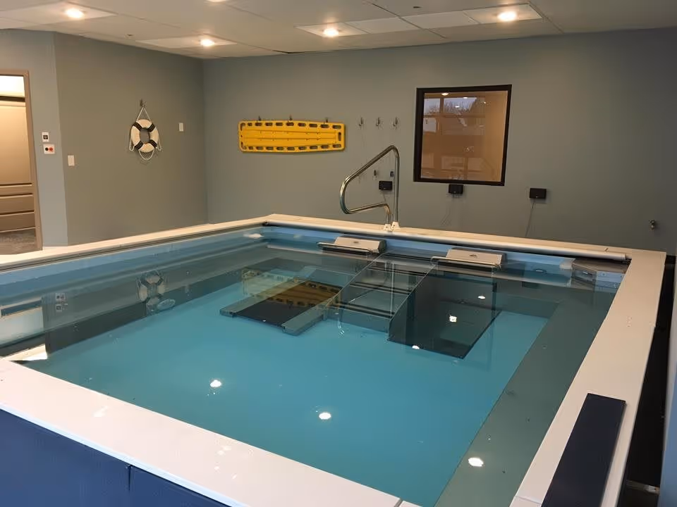 Indoor therapy pool with clear blue water, metal handrails, and underwater treadmill equipment. The room has light blue walls, a window, and safety equipment such as a life ring and a yellow spine board mounted on the wall.