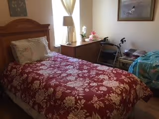 A bedroom with two single beds. The bed in the foreground has a red floral bedspread and a pillow. Between the beds is a wooden nightstand with a lamp, a small plant, and some decorative items. A walker is positioned near the far bed, which has a blue blanket. A framed picture hangs on the wall above the far bed.