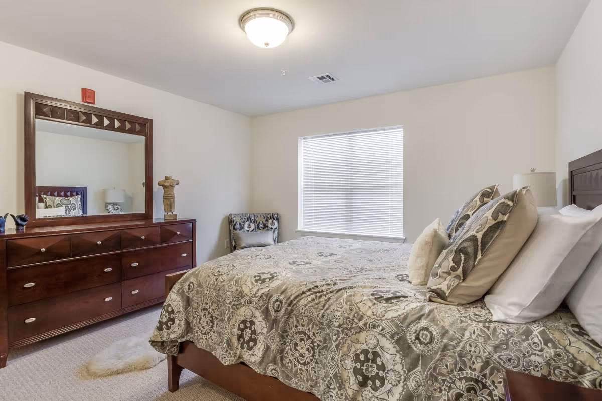 A bedroom with a large bed covered in a patterned bedspread and multiple pillows. There is a wooden dresser with a large mirror above it on the left side of the room, a decorative sculpture on the dresser, and a patterned chair next to a window with closed blinds. The walls are painted white and the room is well-lit with a ceiling light.