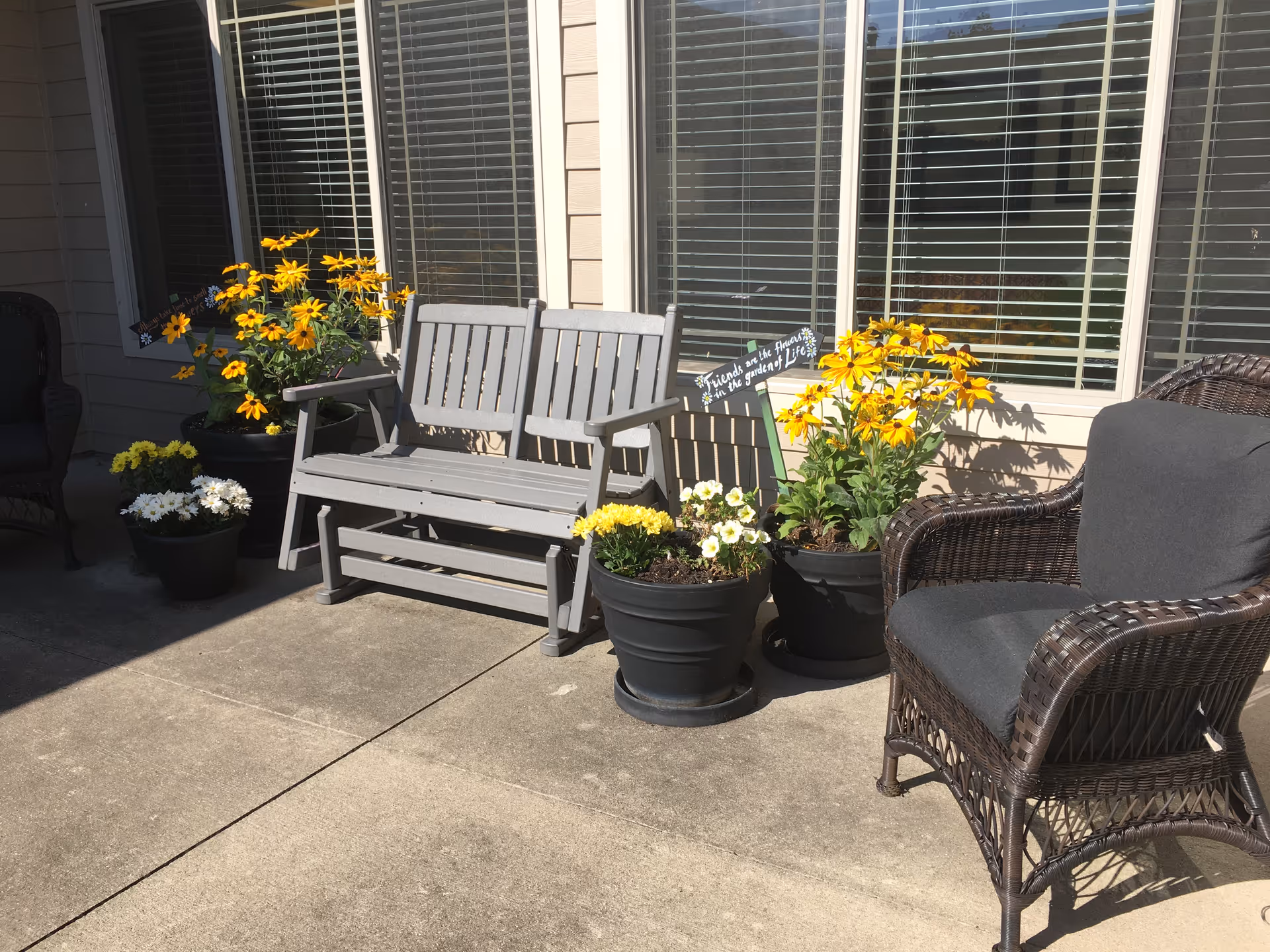 A sunlit outdoor patio with a gray wooden bench, wicker chair, and several pots of yellow and white flowers in front of building windows.