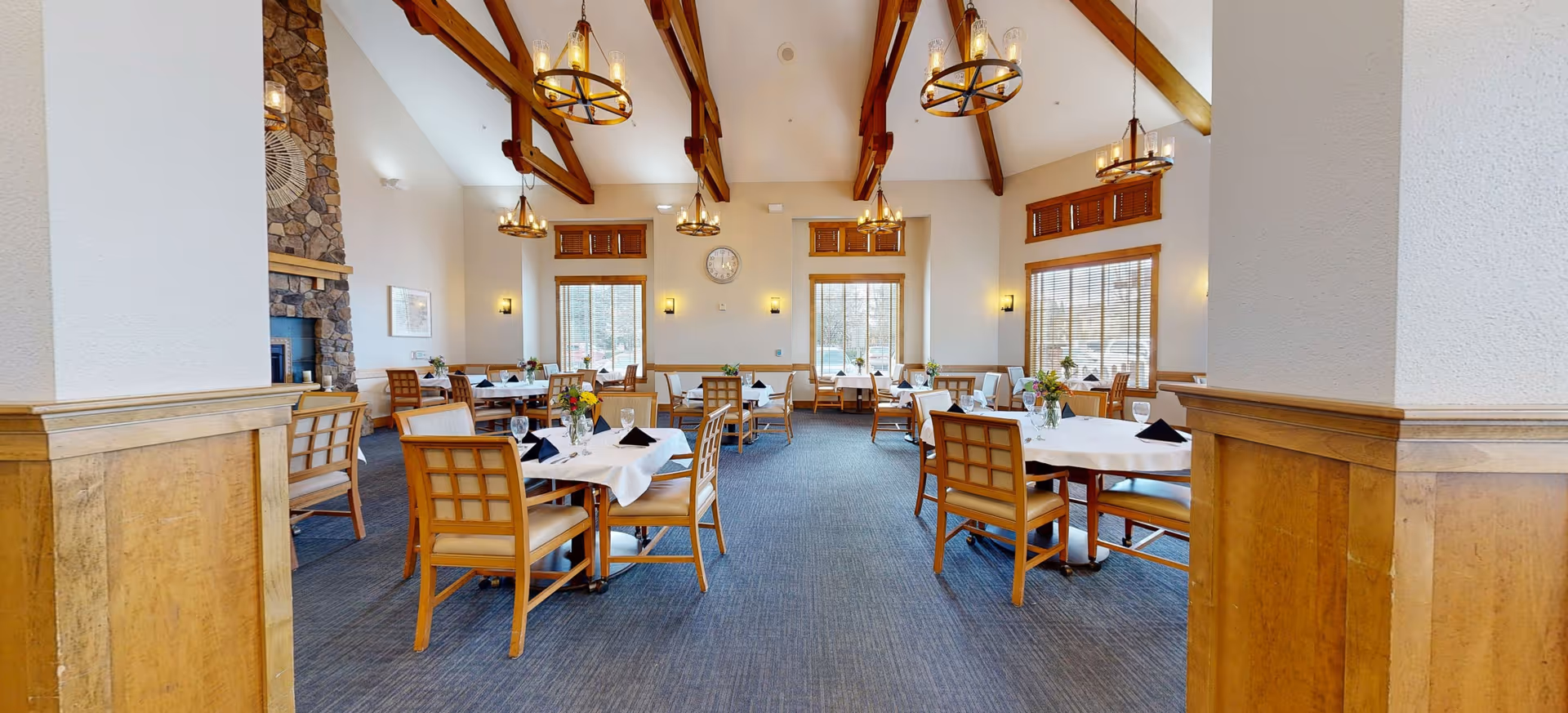 A spacious dining room with multiple round tables covered with white tablecloths, each set with black napkins and glassware. The room features high vaulted ceilings with exposed wooden beams and hanging chandeliers. Large windows with wooden blinds allow natural light to fill the space. There is a stone fireplace on the left side and wooden paneling on the lower part of the walls.