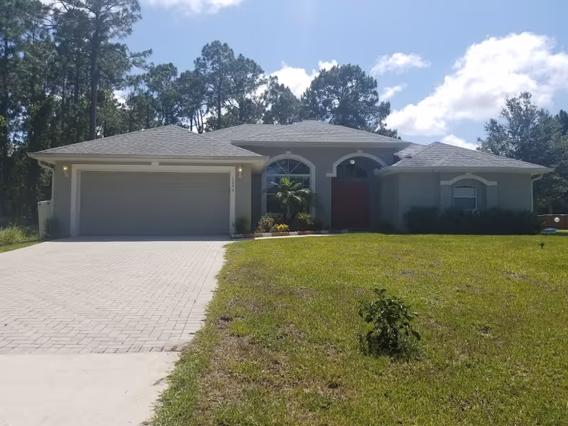 Front view of a single-story house with a two-car garage, a red front door, paved driveway, and a grassy yard with trees behind.