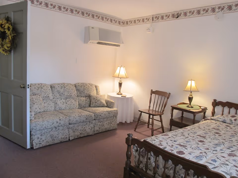 A cozy room with a floral patterned sofa, a wooden bed with a floral quilt, two wooden chairs, two side tables with lamps, and a door with a wreath. The walls are white with a floral border near the ceiling.