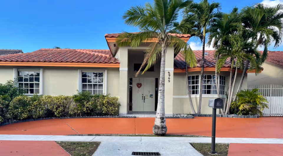 Front exterior view of a single-story residential building with a tiled roof, cream-colored walls, and a central entrance with double doors. There are palm trees and shrubs in front of the building, a mailbox on a post near the sidewalk, and the house number 8969 is visible on a pillar beside the entrance.