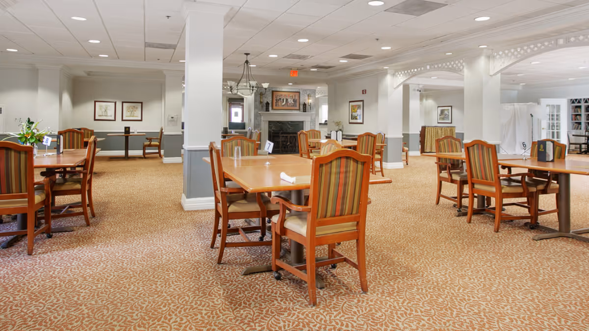 Spacious dining room with wooden tables and striped upholstered chairs arranged on patterned carpet and a fireplace in the background.