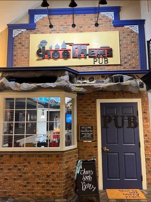 Entrance to a pub named 'Choo Choo's Pub' with a decorative sign featuring a train design above the door. The pub has a brick exterior with a large window and a purple door labeled 'PUB'. There is a small chalkboard sign outside that says 'Cheers and Beers' and a welcome sign with the pub's hours. A doormat in front of the door reads 'this is a happy place.'