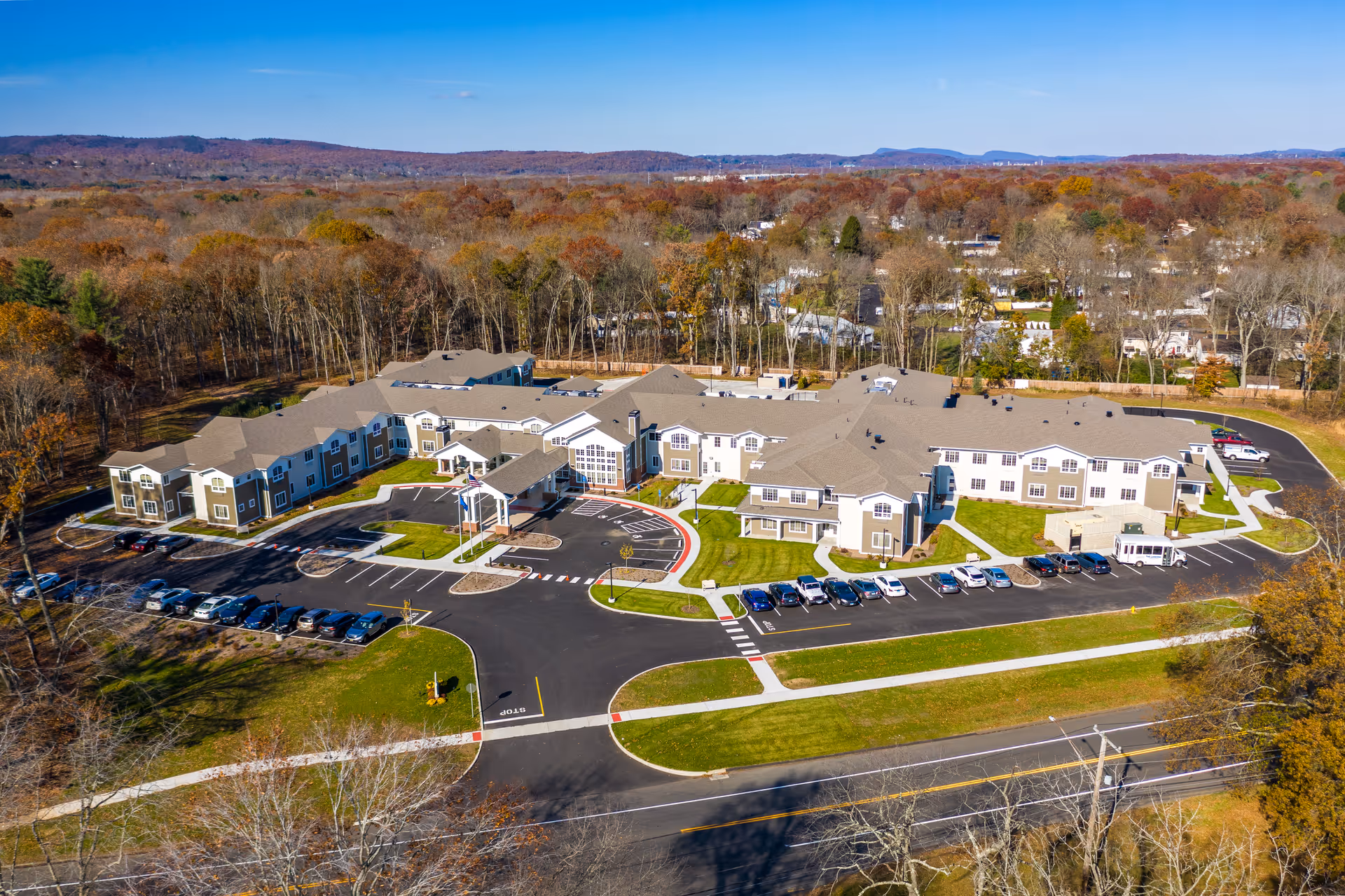 Aerial view of The Landing of North Haven senior living facility showing a large, multi-wing building surrounded by parking lots, green lawns, and trees with autumn foliage. The facility is located near a road and is set against a backdrop of wooded hills and a clear blue sky.