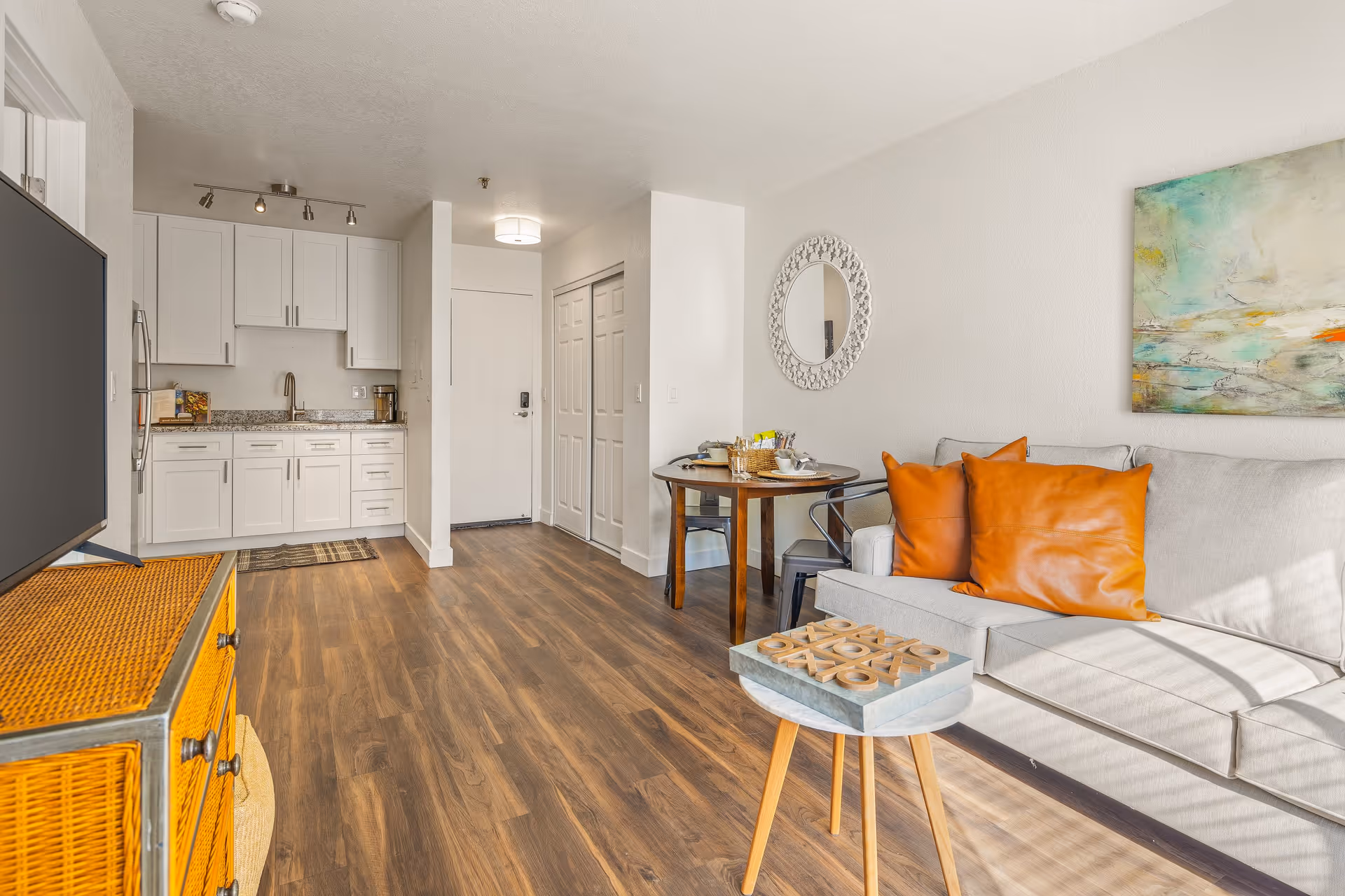A bright and modern living space featuring a small kitchen with white cabinets and granite countertops, a round wooden dining table with two chairs, a light gray sofa with two orange pillows, a small round coffee table with a tic-tac-toe game, a flat-screen TV on a wicker cabinet, a decorative round mirror on the wall, and a colorful abstract painting. The floor is wood, and sunlight streams in from the right side.
