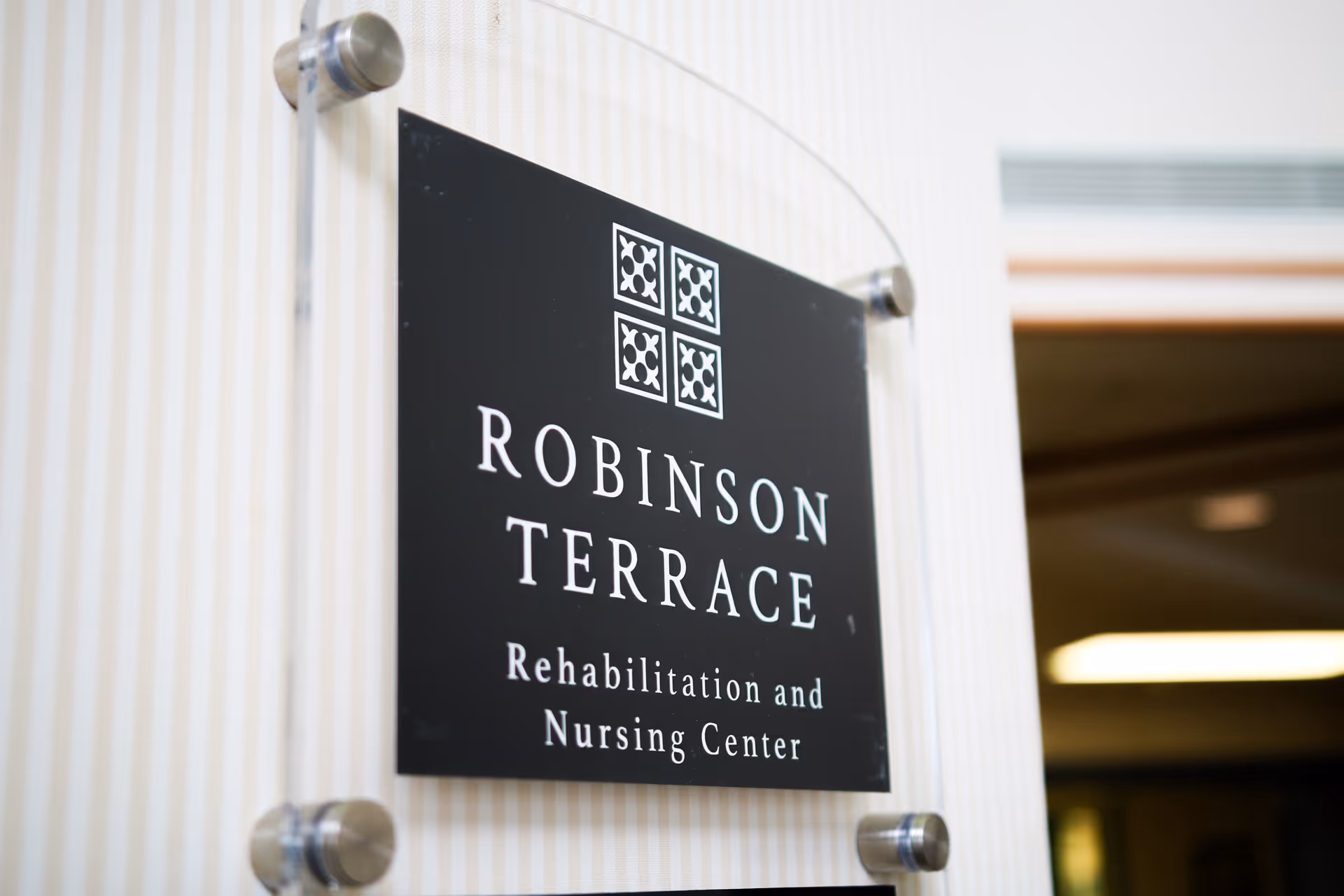 A close-up view of a black sign mounted on a wall with silver standoffs. The sign reads 'ROBINSON TERRACE Rehabilitation and Nursing Center' with a decorative logo above the text. The background wall has light vertical stripes, and part of an interior space with ceiling lights is visible in the background.