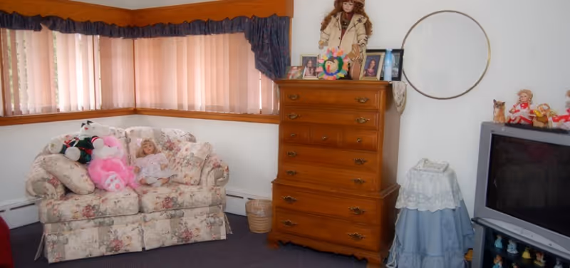 A cozy room with a floral patterned loveseat adorned with stuffed animals and a doll. Next to it is a tall wooden chest of drawers with framed photos and a doll on top. A small round table covered with a blue and white cloth holds a telephone. A TV sits on a stand with small figurines underneath. The room has carpeted flooring, a corner window with vertical blinds and a dark valance, and a circular mirror on the wall.