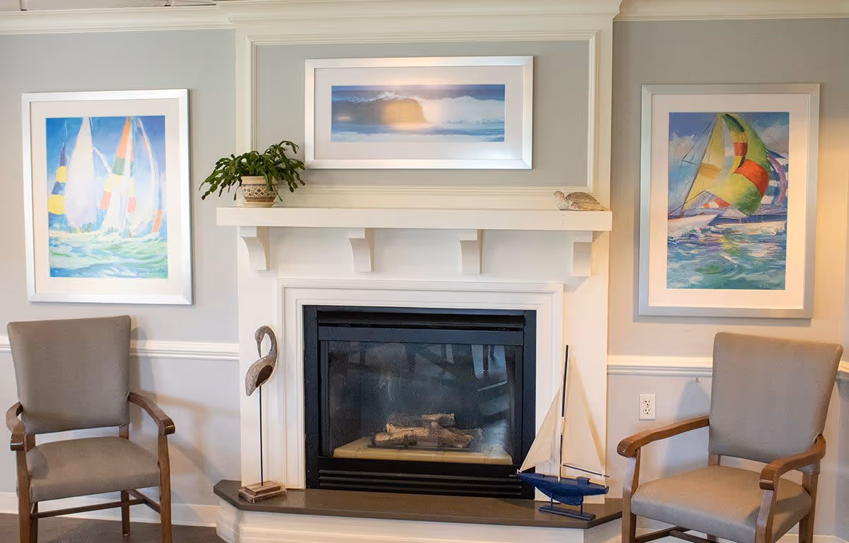 A cozy interior seating area featuring a white fireplace with a black insert, flanked by two beige armchairs with wooden armrests. Above the fireplace are three framed paintings depicting sailboats and ocean scenes. Decorative items including a potted plant, a small sculpture, and a model sailboat are placed on the mantel and hearth.