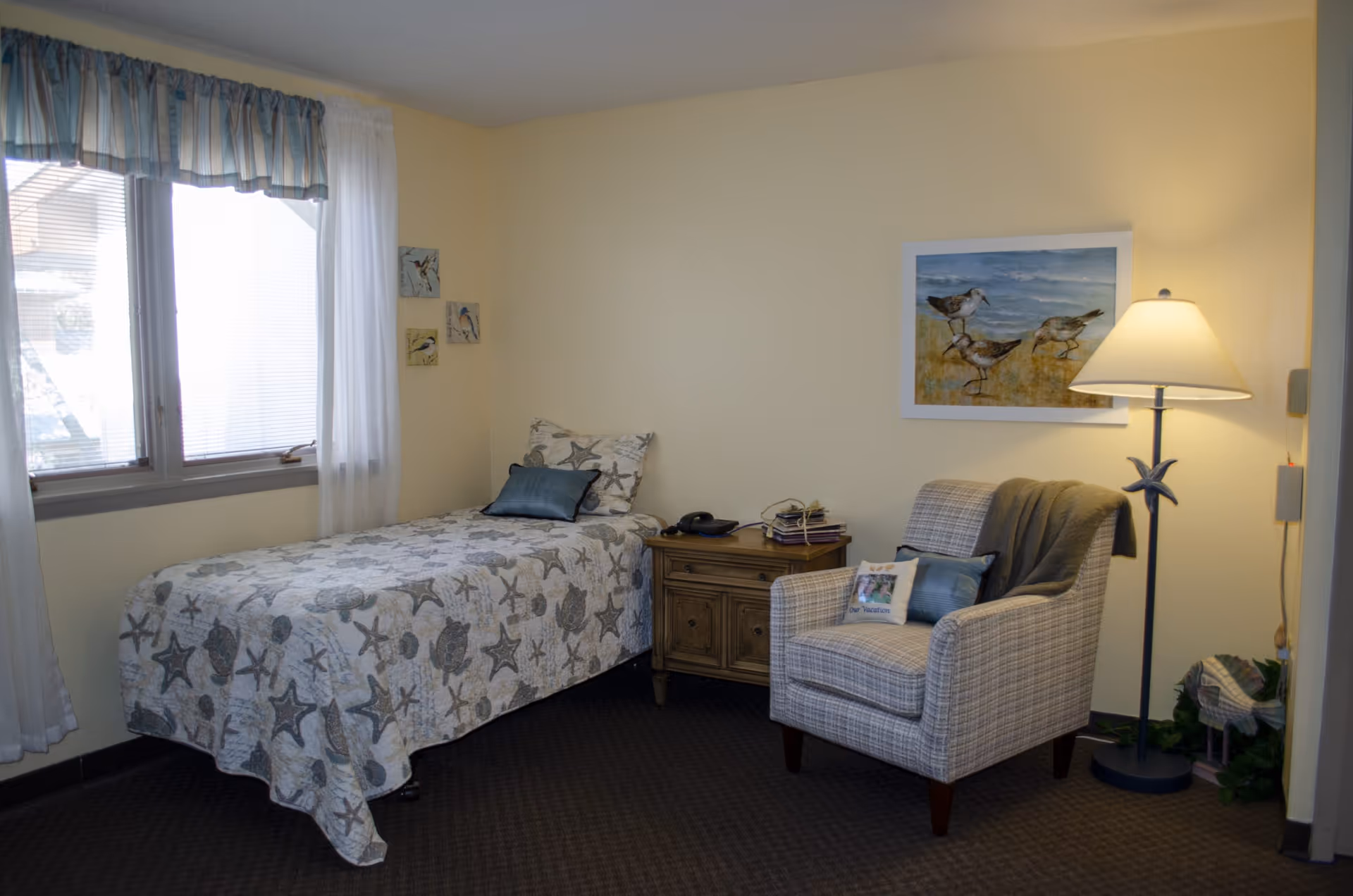 A cozy assisted-living bedroom with a single bed, bedside table, upholstered armchair, and floor lamp.