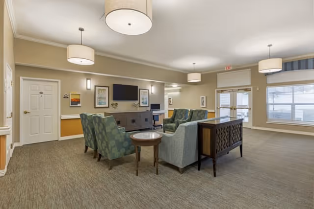 Spacious communal living room with armchairs and sofas arranged around a coffee table facing a wall-mounted TV, pendant lights, and large windows.