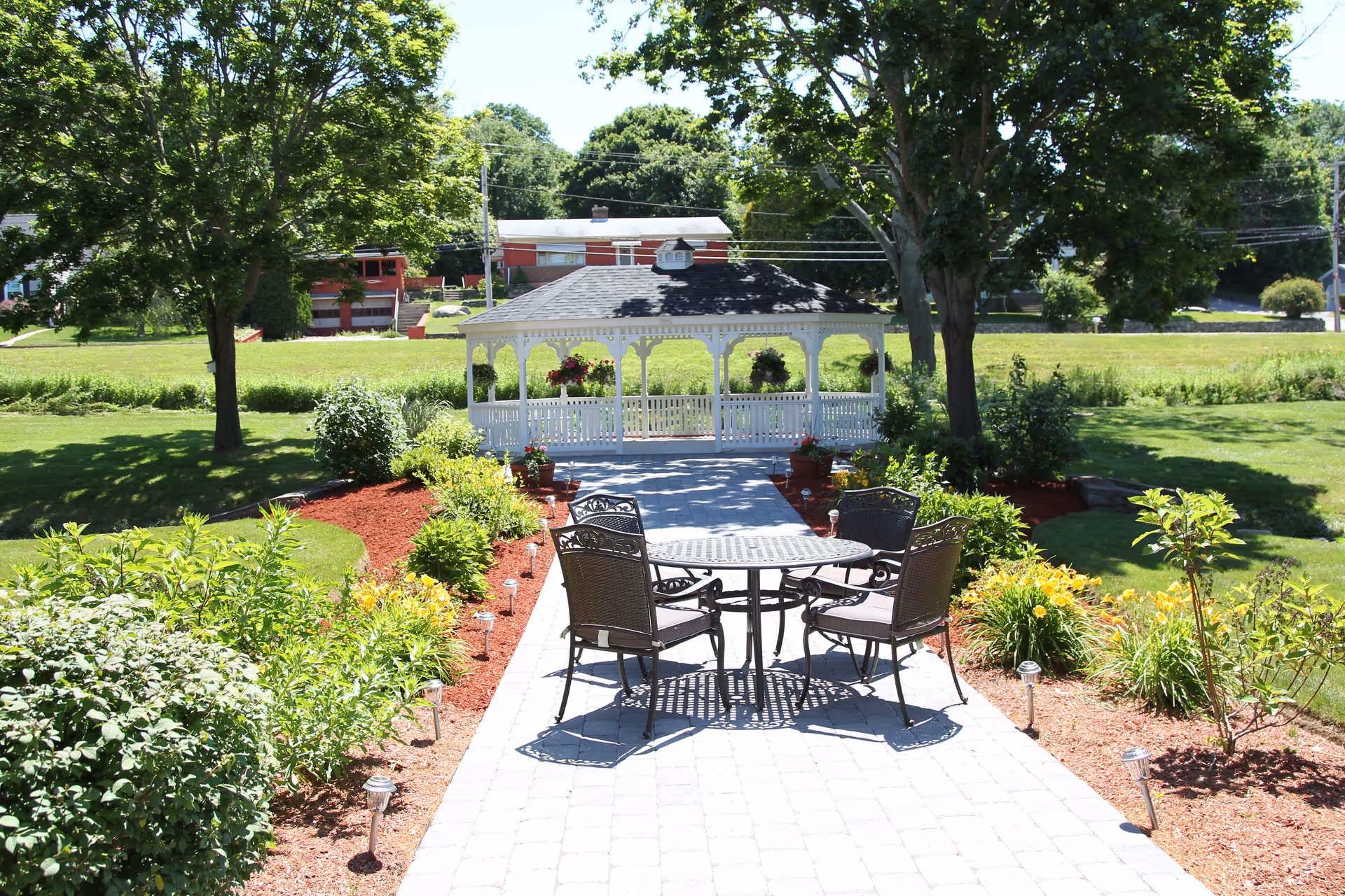 Paved garden path with a round table and chairs leading to a white gazebo amid lawns, trees, and flowerbeds.