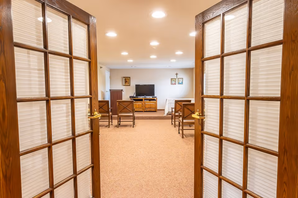 View through open wooden double doors with glass panes into a small chapel or meeting room with carpeted floor, several wooden chairs arranged in rows facing a TV on a wooden stand, and religious pictures and a cross on the far wall.