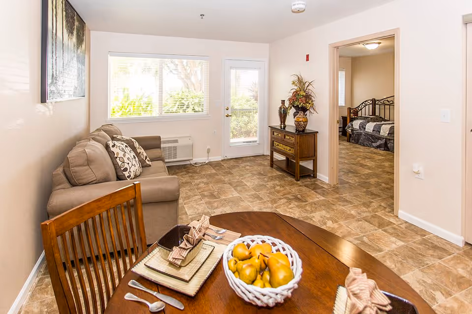 A bright living area in a retirement community apartment featuring a beige sofa with patterned cushions, a wooden dining table set with plates, utensils, and a basket of fruit. The room has tiled flooring, a window with blinds, a door leading outside, and a small wooden cabinet with decorative vases and a flower arrangement. An adjoining bedroom with a bed and metal headboard is visible through an open doorway.